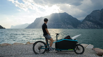 Un hombre está de pie con su bicicleta de carga, a la orilla del lago. Contempla las montañas al fondo.