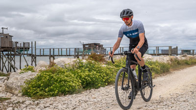 Se ve a un hombre con ropa de ciclista. Va en su bicicleta gravel por un camino de playa.