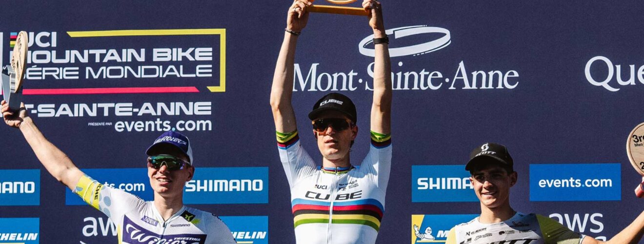 Finn Treudler celebra en la final de temporada del Cube Factory Racing 2025 en Mont-Sainte-Anne, sosteniendo el trofeo en sus manos, flanqueado por los ciclistas que ocuparon el segundo y tercer lugar en el podio.